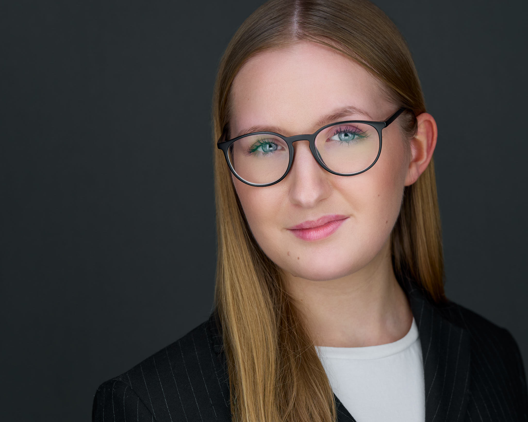 Business-Headshot einer jungen Frau mit Brille und Nadelstreifen-Blazer vor dunkelgrauem Hintergrund, fotografiert von Christian Volk